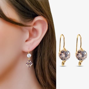 May include: Gold-tone drop earrings with clear and purple-toned crystals. One earring features a round crystal and a square-shaped crystal, while the other earring has a large, round crystal. The earrings are displayed on a light background.
