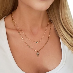May include: Three gold necklaces layered on a person's neck. The top necklace is a delicate chain with a small pendant that says "222". The middle necklace is a slightly thicker chain. The bottom necklace is a thicker chain with a small gold cross pendant. Shown top to bottom: Cressida heart chain, Whitney 222, Celeste chain, Cari cross 16mm.