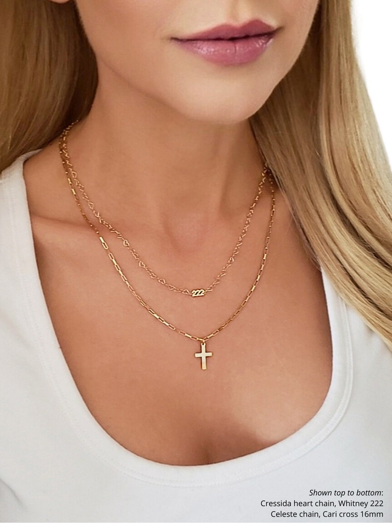 May include: Three gold necklaces layered on a person's neck. The top necklace is a delicate chain with a small pendant that says "222". The middle necklace is a slightly thicker chain. The bottom necklace is a thicker chain with a small gold cross pendant. Shown top to bottom: Cressida heart chain, Whitney 222, Celeste chain, Cari cross 16mm.