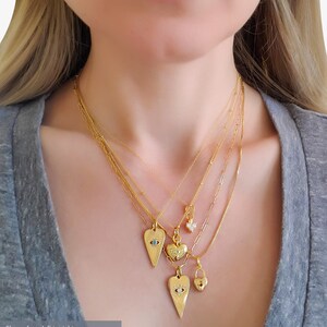 May include: Five gold necklaces with different heart-shaped pendants. The pendants feature a turquoise stone, a heart, a clear stone, a lock, and an evil eye. The necklaces are layered on a person's neck.