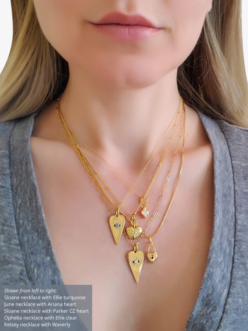 May include: Five gold necklaces with different heart-shaped pendants. The pendants feature a turquoise stone, a heart, a clear stone, a lock, and an evil eye. The necklaces are layered on a person's neck.