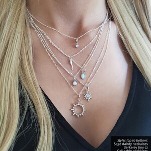 May include: A layered silver necklace set featuring five distinct chains with various charms including a tiny CZ stone, a horn, an oval opal, a round opal, a starburst, and a sun pendant. Text reads: Styles top to bottom: Sage dainty neckalces, Berkeley tiny cz, Cali cable necklaces, Adisa horn pendant, Liva opal oval, Faith opal pendant, June beaded necklace, Raina cz star, Jin cz pendant.