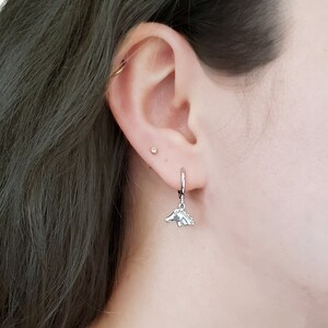 May include: A silver hoop earring with a silver horse charm hanging from it. The earring is worn in the ear of a person with dark hair.