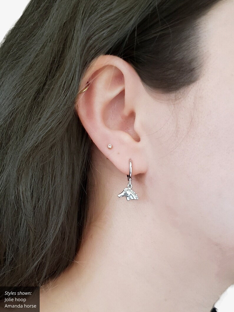 May include: A silver hoop earring with a silver horse charm hanging from it. The earring is worn in the ear of a person with dark hair.