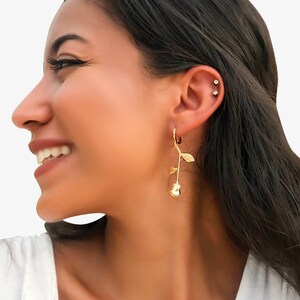 May include: A pair of gold hoop earrings with a rose charm dangling from each hoop. The earrings are shown on a person's ear.
