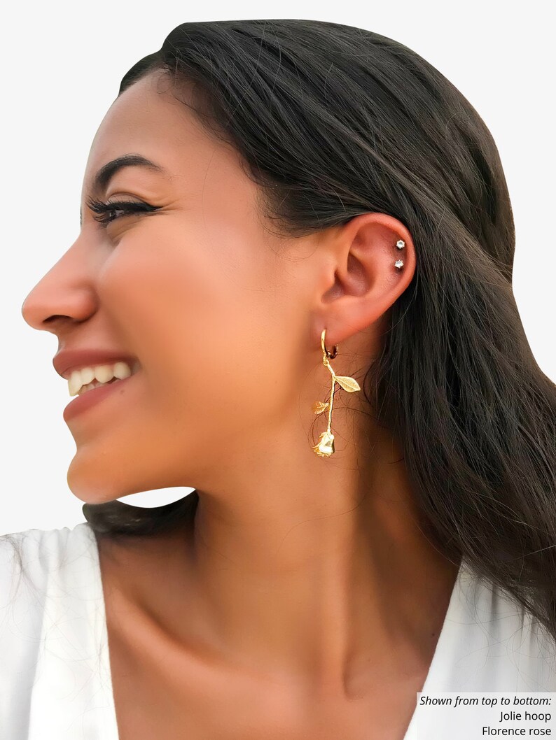 May include: A pair of gold hoop earrings with a rose charm dangling from each hoop. The earrings are shown on a person's ear.