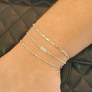 May include: Three silver chain bracelets stacked on a wrist. The top bracelet is a paperclip chain, the middle bracelet is a delicate chain with a charm that says "555", and the bottom bracelet is a bar chain.