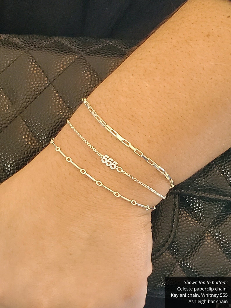 May include: Three silver chain bracelets stacked on a wrist. The top bracelet is a paperclip chain, the middle bracelet is a delicate chain with a charm that says "555", and the bottom bracelet is a bar chain.