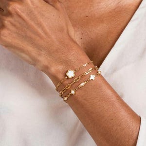 K&ouml;nnte beinhalten: Goldfarbene Armb&auml;nder, die an einem Handgelenk getragen werden. Die Armb&auml;nder umfassen einen Kleeblatt-Charm, eine Kette mit ovalen Gliedern und eine Kette mit kleinen, runden Charms. Der Text im Bild listet die Stile auf: Thalia flower connector, Stevy krinkle chain, Steff sequin 4mm chain, und Tinsley clover chain.