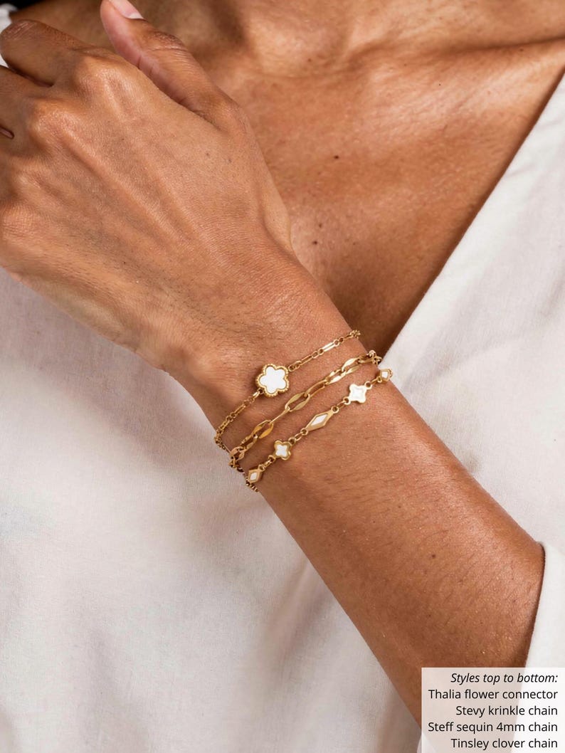 K&ouml;nnte beinhalten: Goldfarbene Armb&auml;nder, die an einem Handgelenk getragen werden. Die Armb&auml;nder umfassen einen Kleeblatt-Charm, eine Kette mit ovalen Gliedern und eine Kette mit kleinen, runden Charms. Der Text im Bild listet die Stile auf: Thalia flower connector, Stevy krinkle chain, Steff sequin 4mm chain, und Tinsley clover chain.