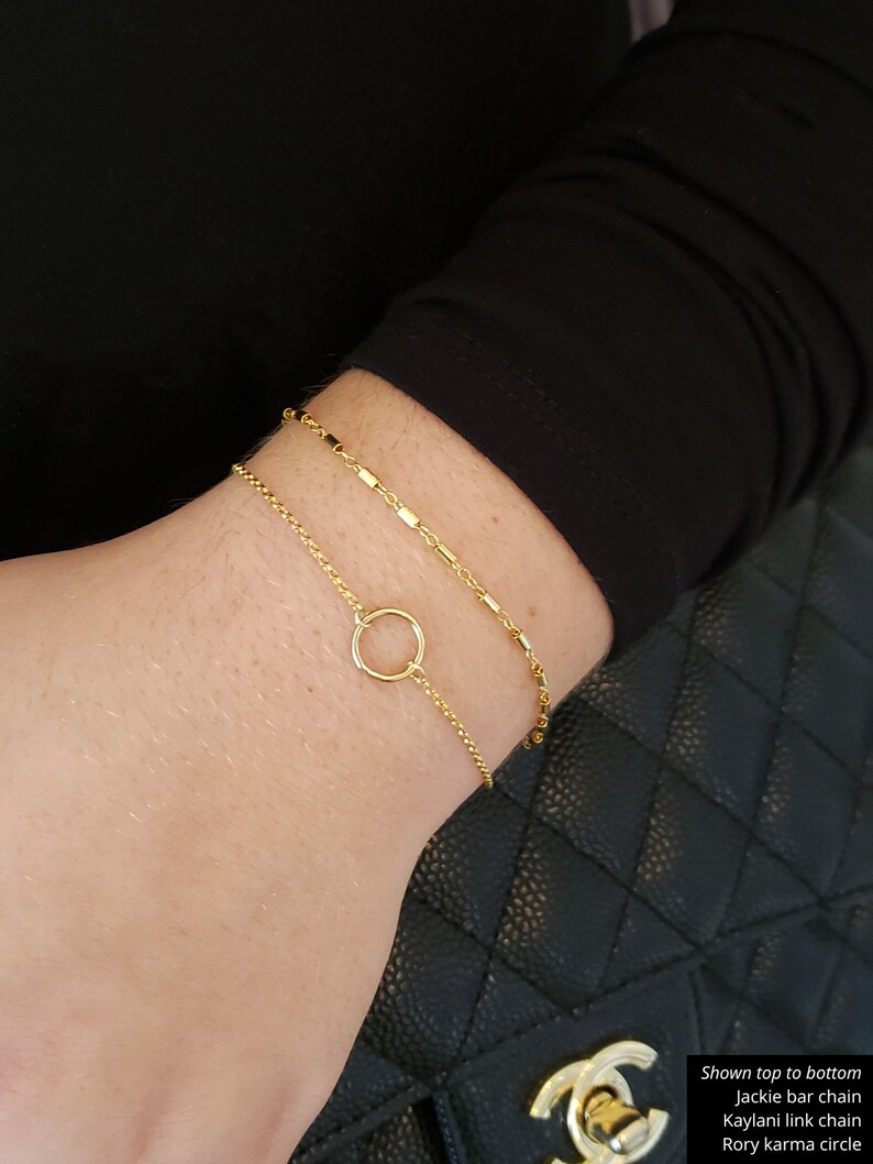 May include: Three gold bracelets stacked on a wrist. The top bracelet is a delicate chain with small square links. The middle bracelet is a simple chain with round links. The bottom bracelet is a circle with a small gap in the circle.