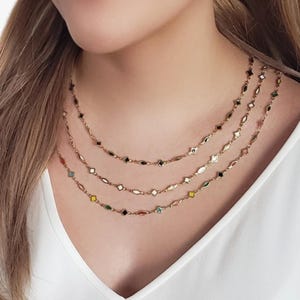 K&ouml;nnte beinhalten: Eine Frau tr&auml;gt drei feine goldene Halsketten. Jede Halskette hat kleine, farbenfrohe, geometrische Anh&auml;nger. Die Anh&auml;nger umfassen Formen wie Diamanten, Ovale und Sterne, mit Farben wie Schwarz, Wei&szlig;, Rot und Gr&uuml;n.
