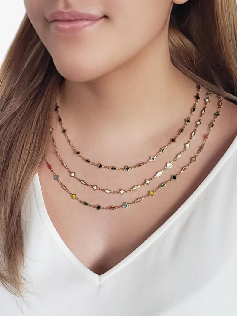 K&ouml;nnte beinhalten: Eine Frau tr&auml;gt drei feine goldene Halsketten. Jede Halskette hat kleine, farbenfrohe, geometrische Anh&auml;nger. Die Anh&auml;nger umfassen Formen wie Diamanten, Ovale und Sterne, mit Farben wie Schwarz, Wei&szlig;, Rot und Gr&uuml;n.
