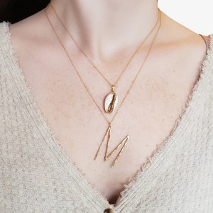 May include: Two gold necklaces with pendants. The top necklace has a small white shell pendant with gold accents. The bottom necklace features a large gold letter "M" pendant. The person is wearing a beige sweater.