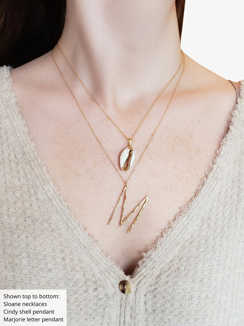 May include: Two gold necklaces with pendants. The top necklace has a small white shell pendant with gold accents. The bottom necklace features a large gold letter "M" pendant. The person is wearing a beige sweater.