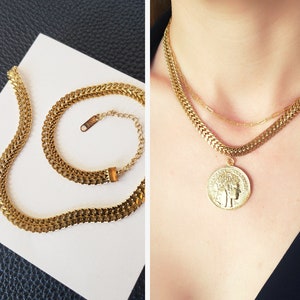 以下が含まれることがあります： 太いチェーンとコインペンダントが特徴のゴールドトーンネックレスセット。 ネックレスは白い面に表示され、人の首の周りに着用されています。 コインペンダントには詳細なレリーフデザインがあります。