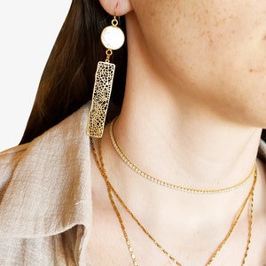 May include: Gold-toned earrings with a white circular accent and a rectangular, openwork design. A gold necklace with a unicorn pendant and other gold necklaces are also visible. The image shows jewelry.