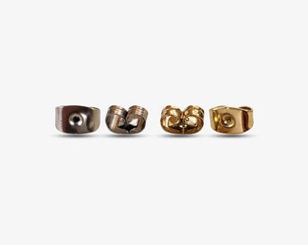 May include: Four earring backs arranged in a horizontal row. The two backs on the left are silver-toned metal, while the two backs on the right are gold-toned metal. Each piece features a circular center hole and curved, wing-like side grips.