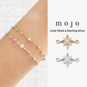 Puede incluir: Dos pulseras delicadas, una chapada en oro y otra de plata de ley, cada una con un dije de estrella con una gema central. La imagen también muestra dos dijes de estrella. Se muestra la palabra "mojo".
