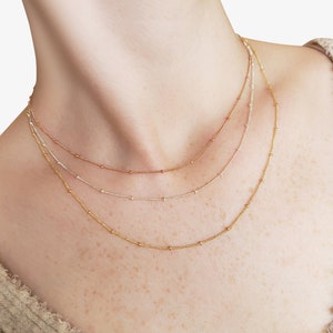 Puede incluir: Tres collares superpuestos con cuentas doradas, plateadas y de oro rosa. Los collares est&aacute;n superpuestos en el cuello de una persona.