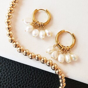 May include: Gold-colored beaded necklace and gold hoop earrings with dangling white pearls. The earrings have a small hoop design and are adorned with multiple teardrop-shaped pearls. The necklace features small, round beads.