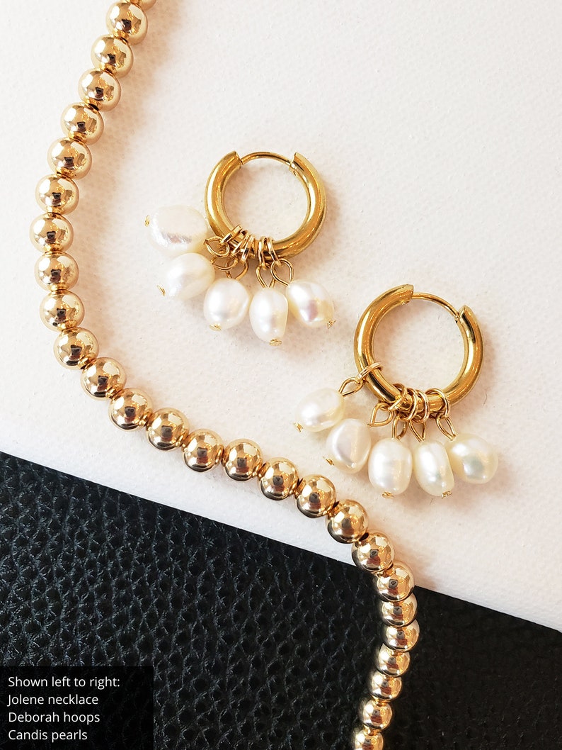 May include: Gold-colored beaded necklace and gold hoop earrings with dangling white pearls. The earrings have a small hoop design and are adorned with multiple teardrop-shaped pearls. The necklace features small, round beads.