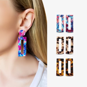 Peut inclure: Une paire de boucles d'oreilles rectangulaires colorées avec un motif marbré. Les boucles d'oreilles sont en acrylique et ont un fermoir doré. Les boucles d'oreilles sont présentées sur l'oreille d'un mannequin.