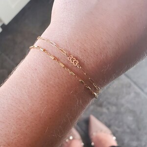 May include: Three gold chain bracelets stacked on a wrist. The top bracelet has a small charm with the number 333. The caption reads "Shown top to bottom: Alexis chain, Whitney 333 Jackie bar chain."