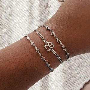 May include: Three silver chain bracelets. The top bracelet features heart charms, the middle one has a paw print charm, and the bottom one has cross charms. The bracelets are worn on a wrist.
