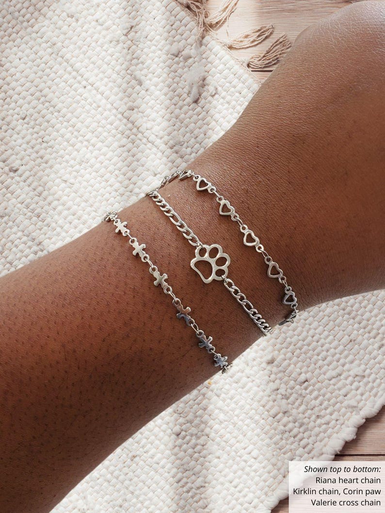 May include: Three silver chain bracelets. The top bracelet features heart charms, the middle one has a paw print charm, and the bottom one has cross charms. The bracelets are worn on a wrist.