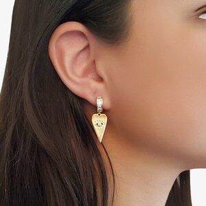 May include: Gold heart-shaped earrings with a small evil eye charm and a clear crystal band.