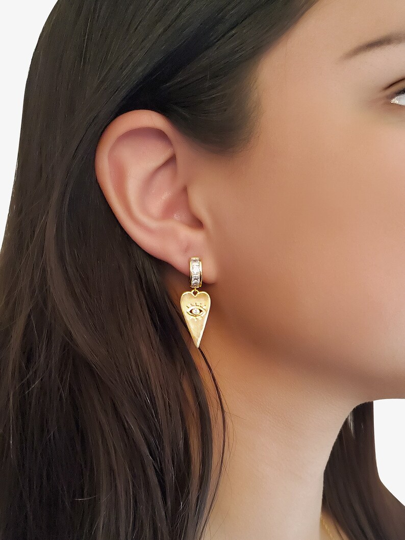 May include: Gold heart-shaped earrings with a small evil eye charm and a clear crystal band.