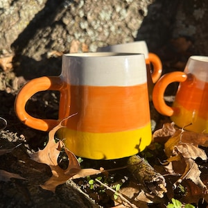 May include: A ceramic mug featuring a horizontal tri-color design with a white top section, an orange middle section, and a yellow bottom section. The mug has a smooth, glossy finish and a curved orange handle attached to the side.