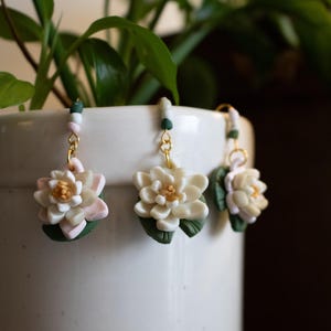 May include: Three handmade polymer clay earrings. Each earring features a white flower with pink accents, a yellow center, and green leaves. The earrings hang from gold-colored hooks with beaded accents. The earrings are displayed in front of a white pot with green foliage.
