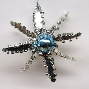 May include: A silver and blue star-shaped Christmas ornament with a shiny blue center. The ornament is made of metal and has a wire loop for hanging.