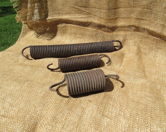 May include: Three rusty metal springs of varying sizes are arranged on a tan burlap surface. The springs have a coiled design with curved hooks at each end. The largest spring is approximately 12 inches long.