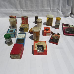 May include: A collection of vintage hardware supplies, including small boxes and containers of tacks, screws, and other fasteners. The items are in various colors, including red, blue, and yellow, and are arranged on a white surface.