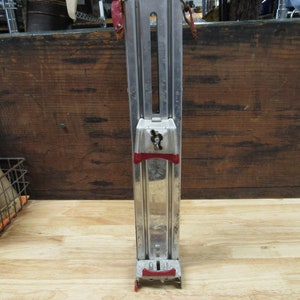 May include: A silver metal can crusher with red plastic handles. The crusher has a lever that is used to crush cans.