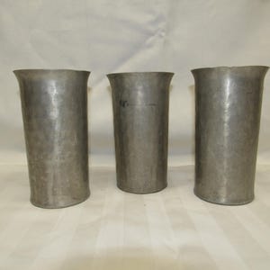 May include: Three antique metal drinking vessels with a hammered texture. The cylindrical cups have a flared rim and a slightly tapered base. The metal has a dull, gray finish, suggesting age and use. The cups are arranged in a row.