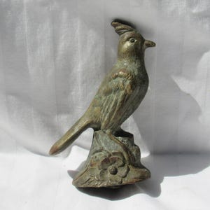 May include: A bronze-toned metal bird figurine featuring a prominent crest on its head, perched atop a base decorated with a sculpted flower. The bird has a textured surface with visible patina, a pointed beak, and a long tail extending backward.