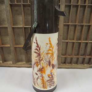 May include: A brown and white vintage thermos with a floral design. The thermos has a handle and a spout.