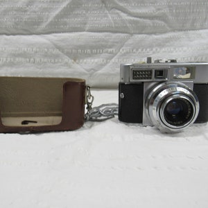 May include: A silver and black 35mm film camera with a leather case and a silver chain. The camera has a lens with a black ring around it and a viewfinder on the top.