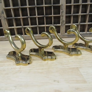May include: Four gold-toned metal hooks with a decorative, ornate design. The hooks are mounted on a wooden surface.