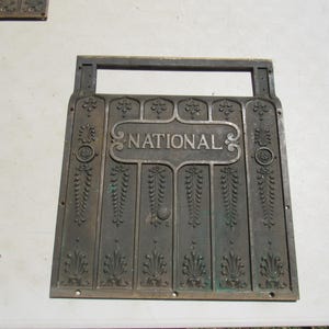 May include: A bronze-colored metal sign with the word "NATIONAL" in a decorative frame. The sign has an ornate design with vertical panels, floral accents, and a handle. The sign is likely an antique or vintage piece.