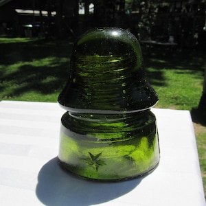 May include: A dark green glass insulator with a star design. The vintage electrical component has a rounded top and a flared base. The glass has a glossy finish and sits on a white surface, with a green grassy background.