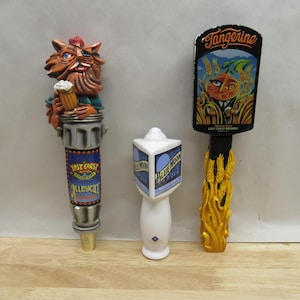 May include: Three beer tap handles. The left handle is a silver and blue handle with a cartoon cat holding a beer mug. The handle has the text "Lost Coast" and "Ale Cat". The middle handle is white with a blue and white label that says "Blue Moon". The right handle is black with a yellow wheat design and a label that says "Tangerine" and "Lost Coast Brewery".
