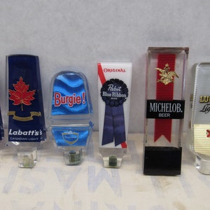 May include: Six different beer tap handles. Two are blue with a red maple leaf and the text "Labatt's Canadian Light". One is blue with a wave pattern and the text "Burgie!". One is white with a blue ribbon and the text "Pabst Blue Ribbon". One is red with a gold eagle and the text "Michelob Beer". One is silver with the text "Lucky Light".