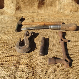 May include: An assortment of vintage metal tools, including a hammer, chisel, hook, hinge, and a unique L-shaped piece. The tools are made of metal and wood, with a rustic, aged appearance. The items are arranged on a burlap surface.