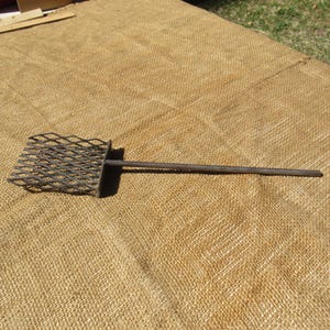 May include: A metal grilling spatula with a diamond-patterned head and a long, straight handle. The spatula is made of dark metal and is designed for use with a grill or barbecue. The tool is lying on a tan, textured surface.