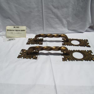 May include: Two brass-colored metal door handles with a twisted bar design. Each handle features rectangular backplates with ornate, textured detailing and a circular opening. A small paper tag labeled NO. 1623 DECORATIVE TWISTED HANDLE is included.